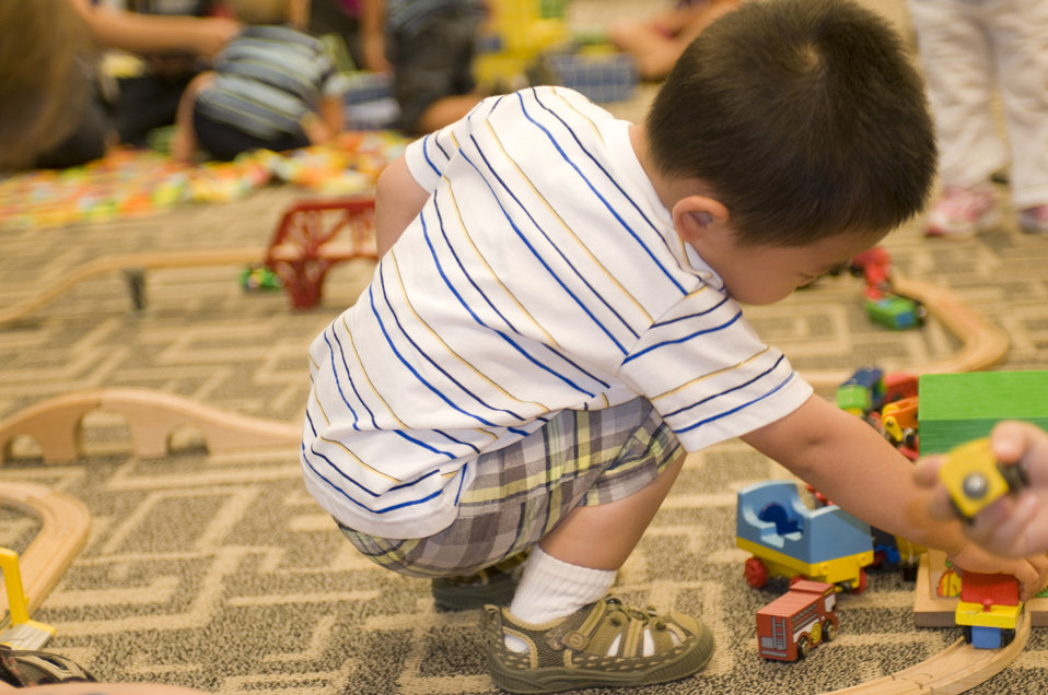 While in a daycare setting, this young boy was entertaining himself by playing with a small wooden toy car set, pushing the little cars alon